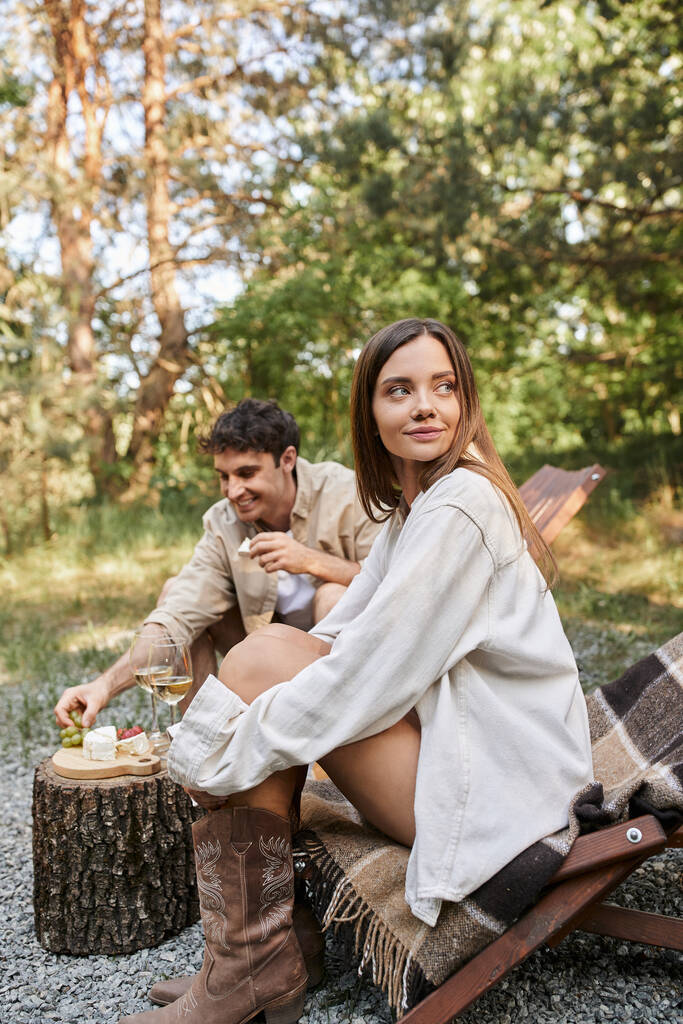 Brunetka kobieta siedzi na leżaku obok chłopaka z winem i jedzeniem podczas pikniku na świeżym powietrzu - Zdjęcie, obraz