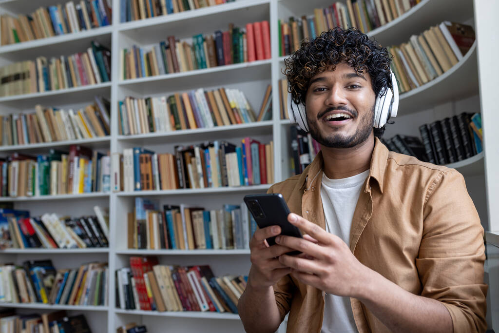 Joven estudiante hispano dentro de la biblioteca pública estudiando, sonriendo y mirando a la cámara, hombre sosteniendo el teléfono, usando la aplicación de estudio, con auriculares entre estantes. - Foto, imagen