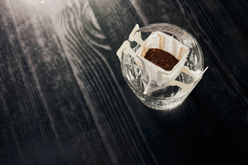bovenaanzicht van aromatische gemalen koffie in papieren filter en kristalglas op zwarte tafel, pour-over brouwsel - Foto, afbeelding