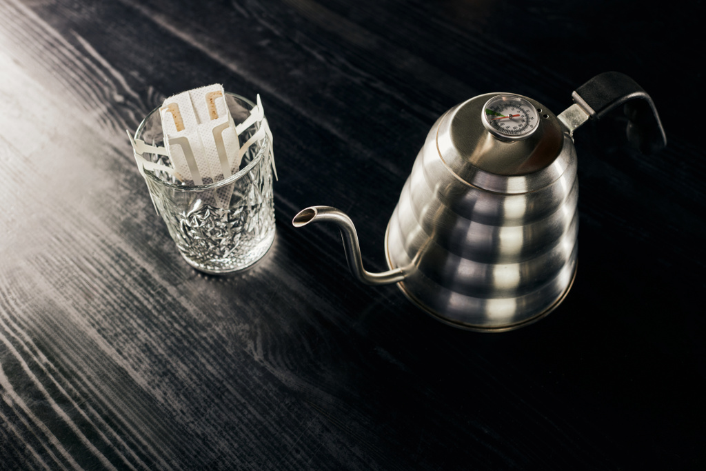 způsob přelévání, kovová kapací konvice, sklo s mletou kávou ve filtračním sáčku na černém stole - Fotografie, Obrázek