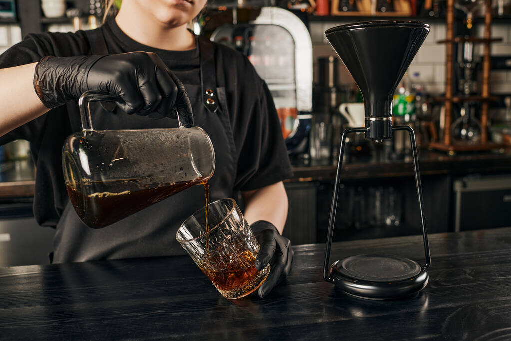 カフェのサイフォンコーヒーメーカー近くのクリスタルガラスに新鮮なエスプレッソを注ぐバリスタの部分的な眺め - 写真・画像
