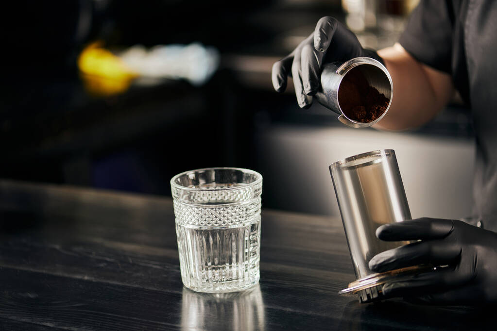 vista parcial de barista en vidrio de látex negro sosteniendo prensa aerodinámica con café cerca de vidrio sobre mesa negra - Foto, imagen