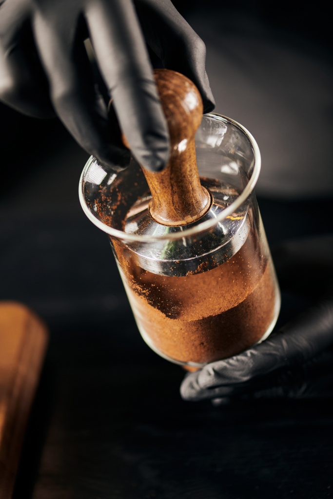 cropped view of barista in black latex gloves pressing ground coffee in cold drip coffee maker in cafe - Photo, Image