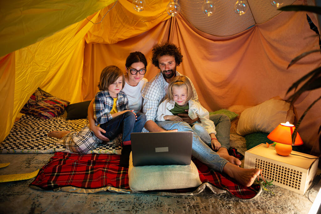 Έννοια του οικογενειακού τρόπου ζωής ευτυχισμένη οικογένεια μαζί βλέποντας ταινία στο φορητό υπολογιστή, ενώ κάθεται μέσα στο χειροποίητο σκηνή στο παιδικό δωμάτιο. - Φωτογραφία, εικόνα