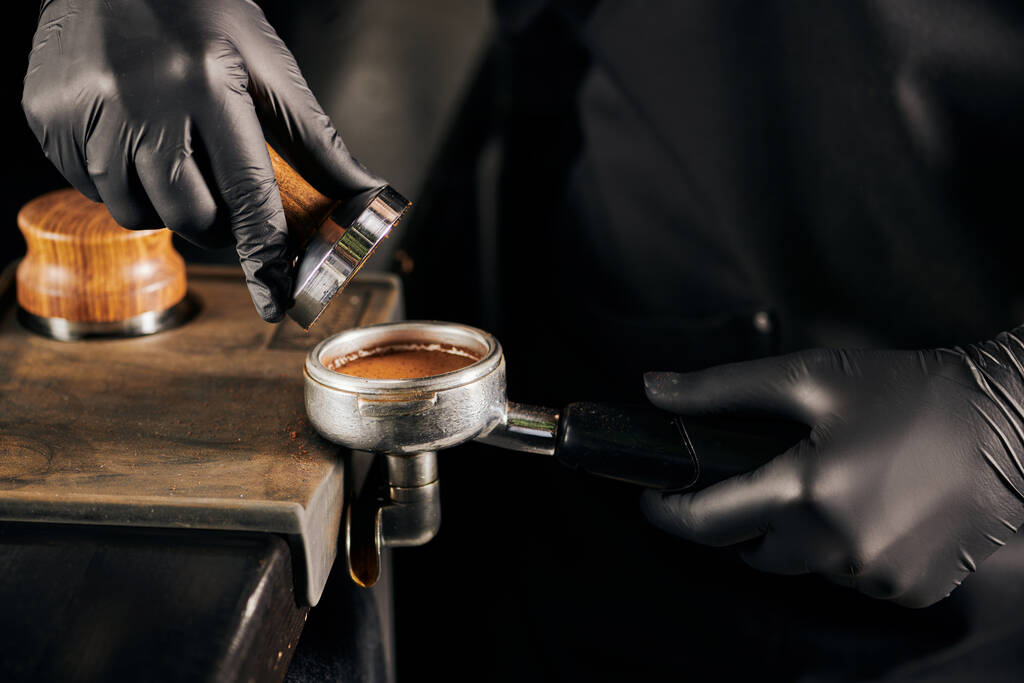 обрезанный вид на бариста в черных латексных перчатках прессовать молотый кофе в портативном фильтре, кафе - Фото, изображение