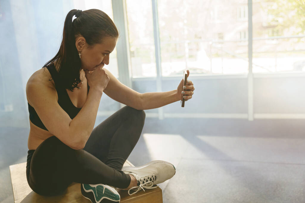 Fit žena ve sportovním oblečení používat telefon během odpočinku v posilovně. Kvalitní fotografie - Fotografie, Obrázek