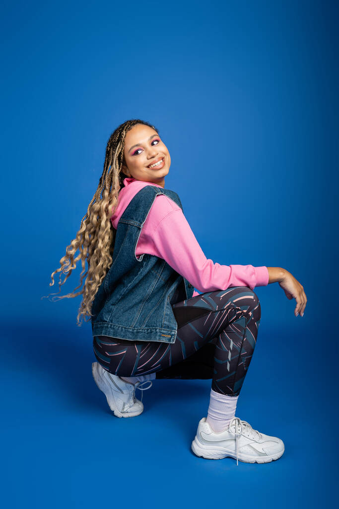 happy african american woman with dreadlocks sitting and posing on blue background, sporty outfit - Photo, Image