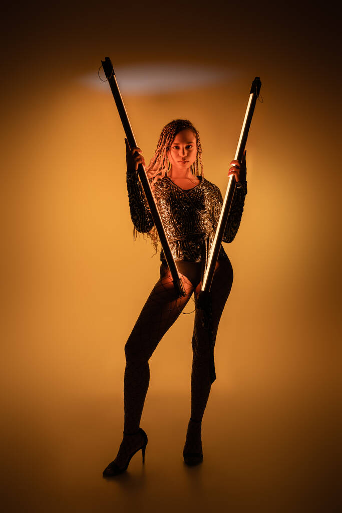 african american woman holding fluorescent lamps on yellow background, high heels and fishnet tights - Photo, Image