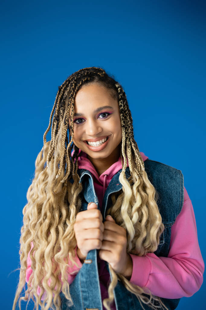 happy african american woman in pink hoodie and denim vest smiling on blue background, dreadlocks - Photo, Image