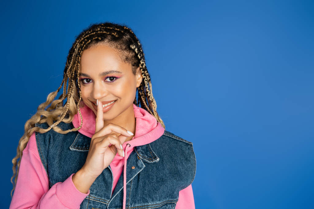 happy african american woman with dreadlocks showing hush sign, blue background, finger near lips - Photo, Image