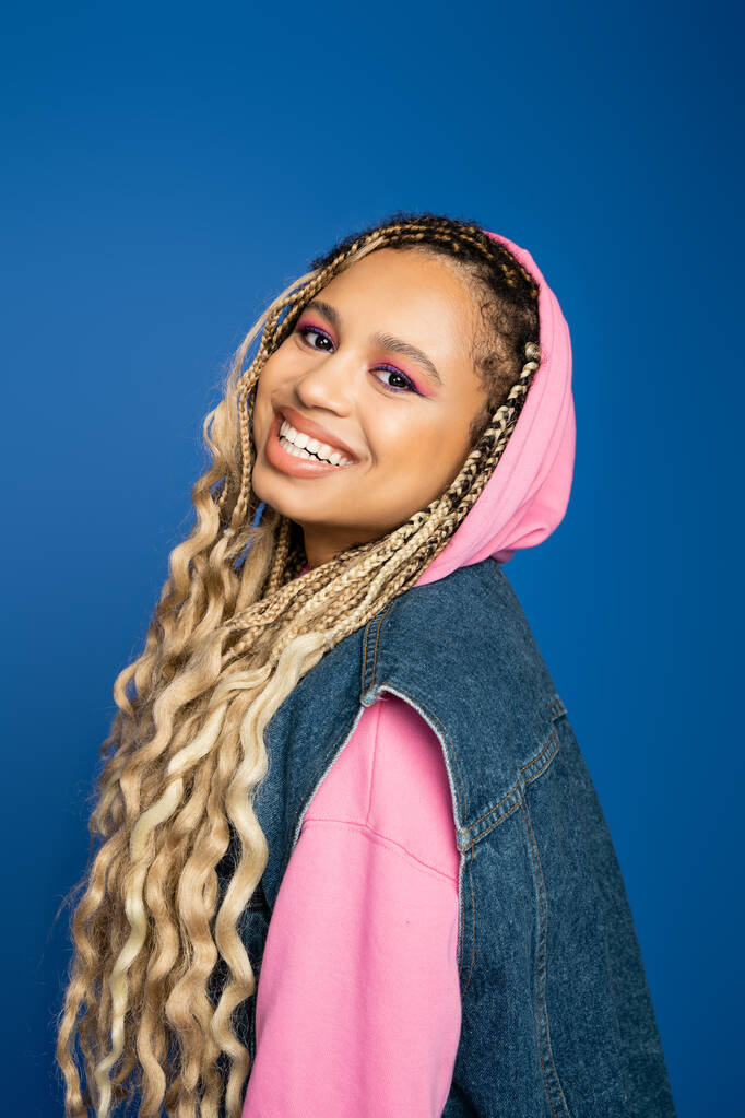 joyful and pretty african american woman with hood on head smiling on blue background, positive - Photo, Image