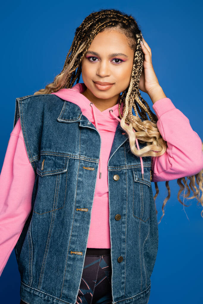dark skinned woman with bold makeup smiling on blue, wearing pink hoodie and denim vest in studio - Photo, Image