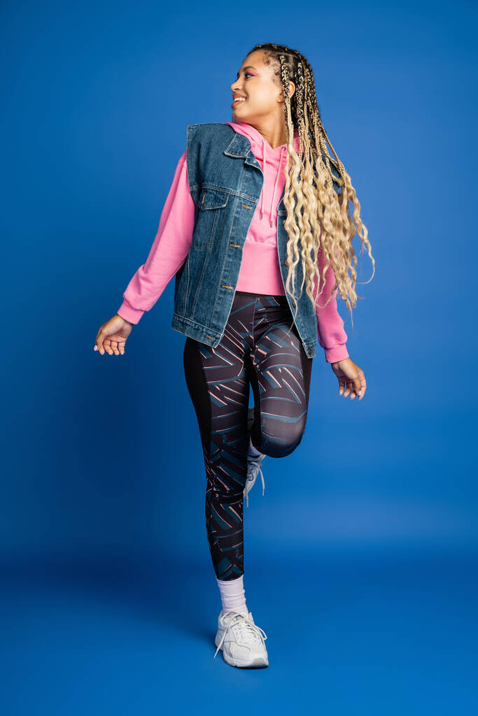 повна довжина, темношкіра жінка в спортивному вбранні, посміхаючись на синьому фоні, позитивний, сміливий макіяж - Фото, зображення