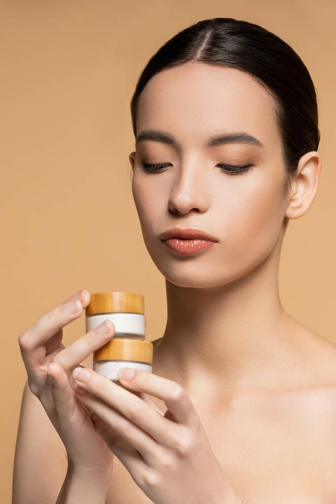 Young asian woman with naked shoulders and perfect skin holding cosmetic cream isolated on beige - Photo, Image
