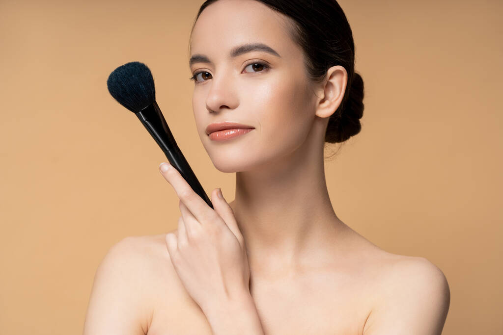 Confident young asian woman with naked shoulders holding makeup brush and standing isolated on beige - Photo, Image