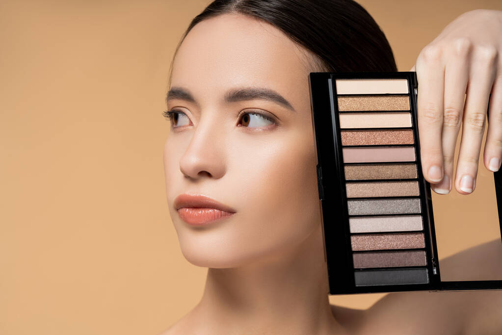 Portrait of young asian woman with natural makeup holding eyeshadow palette isolated on beige - Photo, Image