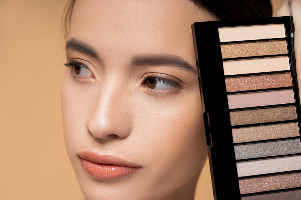 Close up view of young asian woman with natural makeup holding eyeshadow palette isolated on beige - Photo, Image