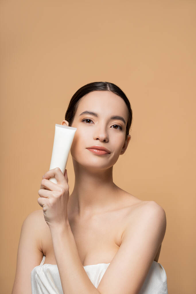 Portrait de jeune modèle asiatique en haut tenant le baume cosmétique et regardant la caméra isolée sur beige - Photo, image