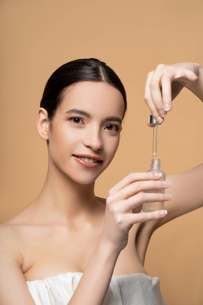 Positive young asian woman with naked shoulders holding cosmetic serum isolated on beige - Photo, Image