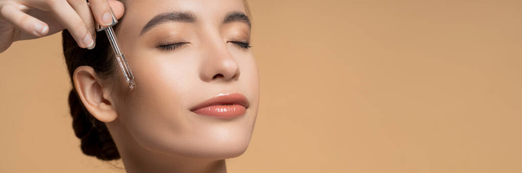 Bella giovane donna asiatica con gli occhi chiusi applicando siero cosmetico isolato sul beige, banner - Foto, immagini