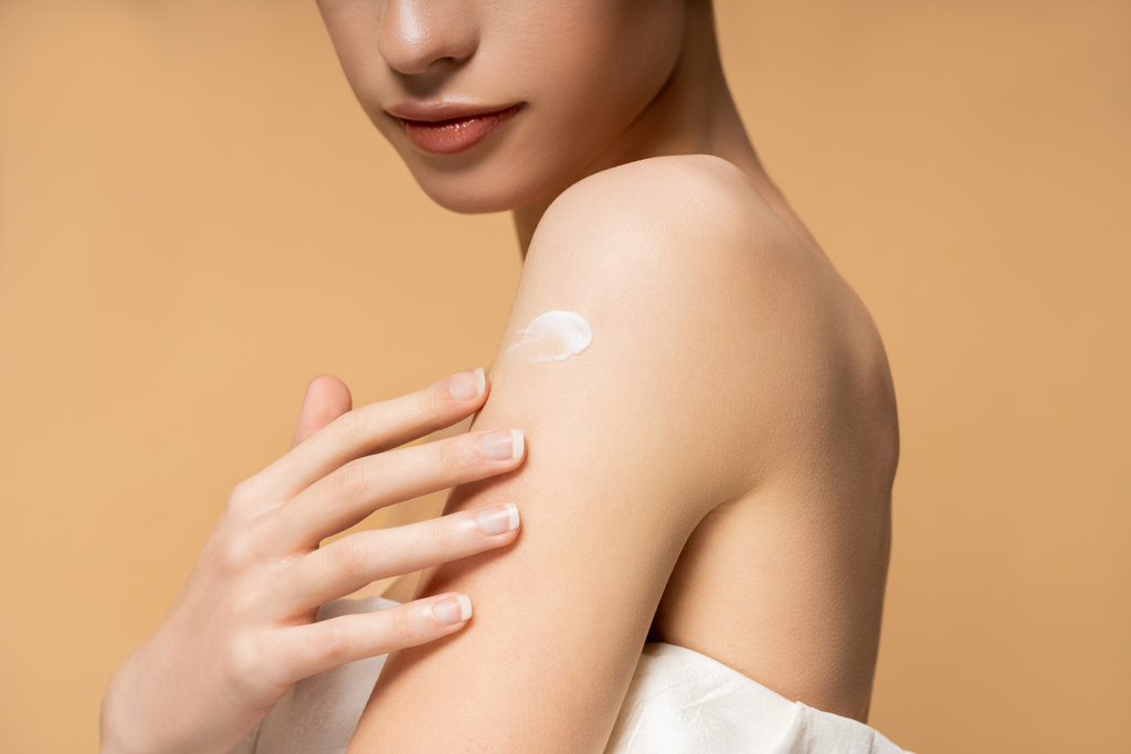 Cropped view of young woman in top with naked shoulder applying body cream isolated on beige - Photo, Image