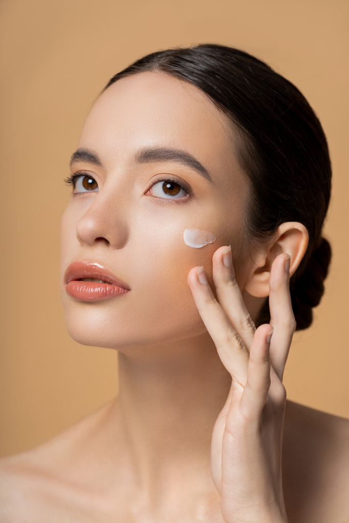 Young asian woman with naked shoulders applying face cream isolated on beige - Photo, Image
