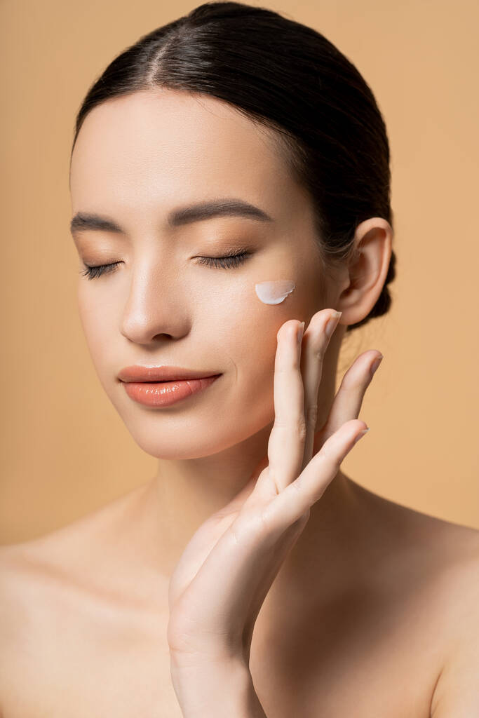 Young asian model with closed eyes applying face cream on cheek isolated on beige - Photo, Image