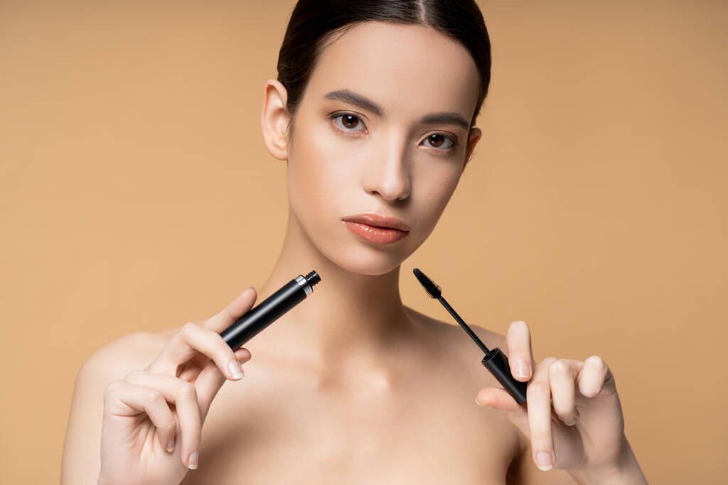 Confident asian woman with naked shoulders holding mascara and applicator isolated on beige - Photo, Image