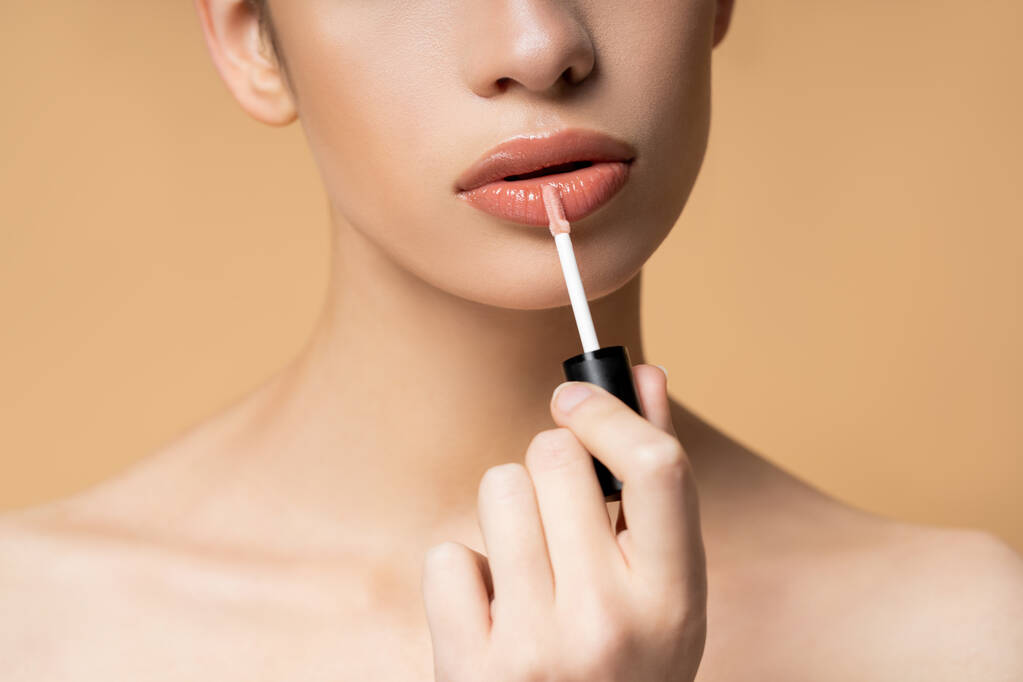 Καλλιεργημένη άποψη του νεαρού μοντέλου με γυμνούς ώμους εφαρμογή lip gloss απομονώνονται σε μπεζ - Φωτογραφία, εικόνα