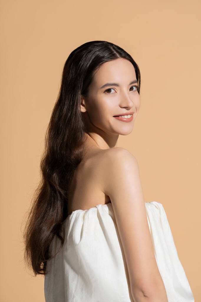 Fröhliches junges asiatisches Model mit langen Haaren schaut in die Kamera, während sie isoliert auf beige steht - Foto, Bild