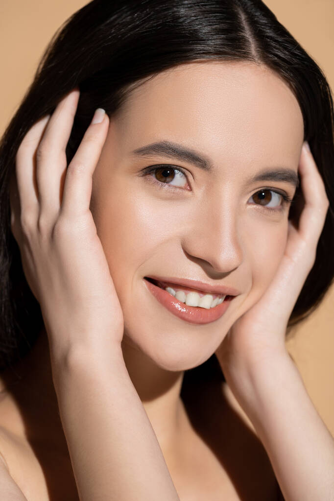 Retrato de modelo asiático positivo con maquillaje natural tocando el cabello aislado en beige - Foto, Imagen