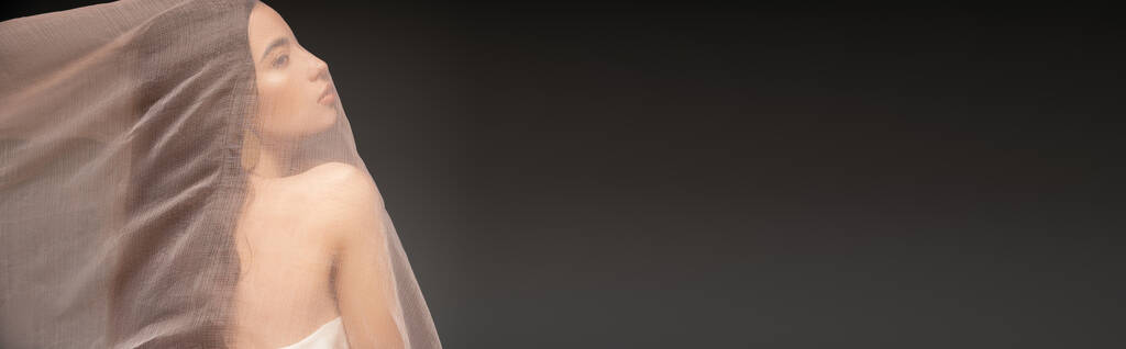 Πλευρική άποψη της νεαρής Ασιάτισσας με γυμνό ώμο που ποζάρει κάτω από ύφασμα απομονωμένο σε μαύρο, λάβαρο - Φωτογραφία, εικόνα