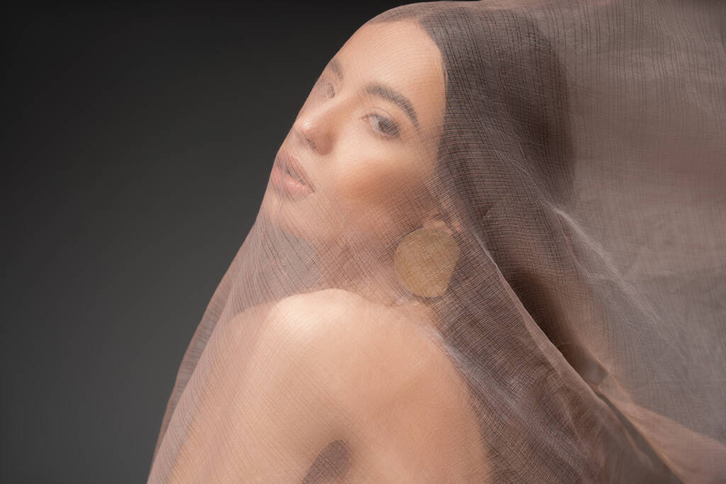 Young asian model with naked shoulder looking at camera under beige cloth isolated on black - Photo, Image