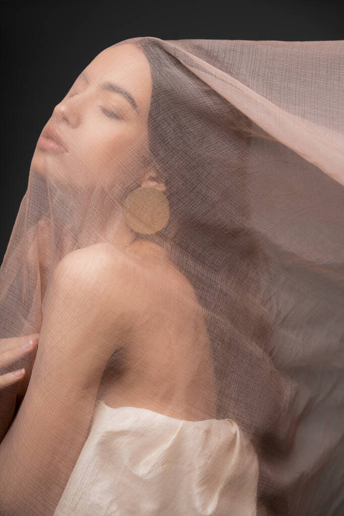 Jolie modèle asiatique en haut et boucle d'oreille posant sous un tissu beige tout en se tenant isolé sur noir - Photo, image
