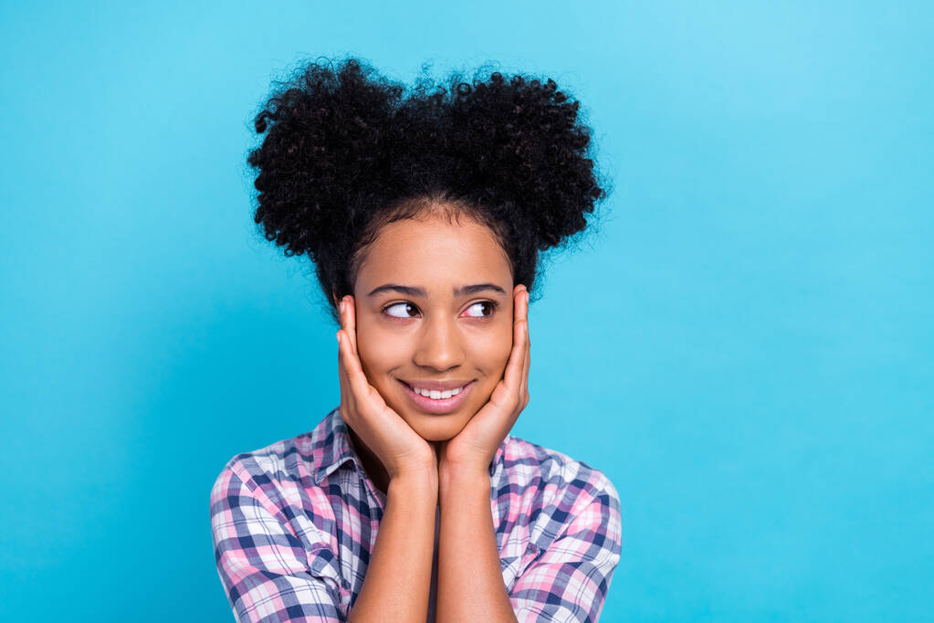 Retrato de optimista adolescente agradable pelo afro camisa a cuadros brazos en los pómulos mirada espacio vacío aislado sobre fondo de color azul. - Foto, imagen