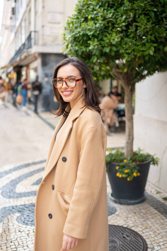 Приємно усміхатися, коли приваблива жінка залишає будинок на зеленому фоні на вулиці європейського міста. Щаслива жінка в окулярах вдягнена стильна траншейна пальто посміхається дивлячись на камеру - Фото, зображення