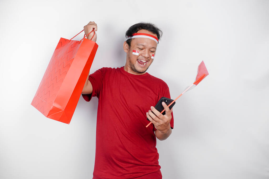 Un atractivo hombre asiático de pie emocionado sosteniendo una bolsa de compras en línea y su teléfono inteligente, estudio de tiro aislado sobre fondo blanco. Concepto del Día de la Independencia de Indonesia - Foto, Imagen