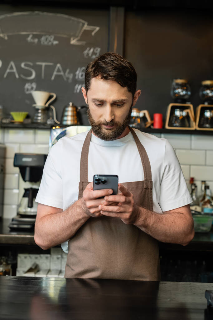 vousatý barista v zástěře pomocí smartphonu při stání v blízkosti baru a práci v kavárně - Fotografie, Obrázek