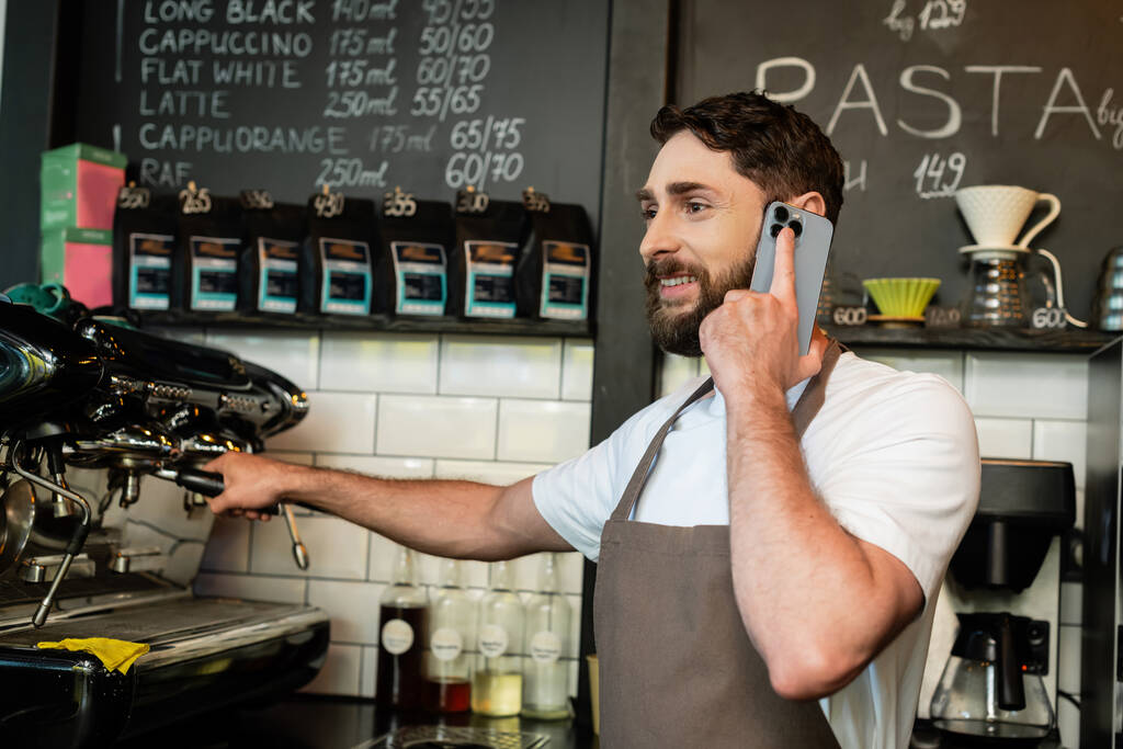 barista sonriente en delantal hablando en el teléfono inteligente mientras trabaja cerca de la máquina de café en la cafetería - Foto, Imagen