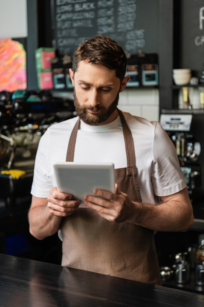 szakállas barista kötényben digitális táblagéppel, miközben dolgozik, és a kávézó bár közelében áll - Fotó, kép