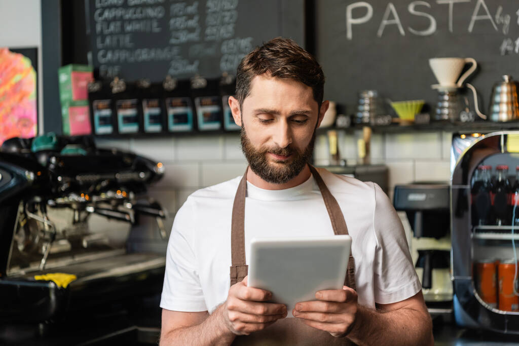 barista in schort met behulp van digitale tablet tijdens het werken en staan in de buurt bar in coffeeshop - Foto, afbeelding