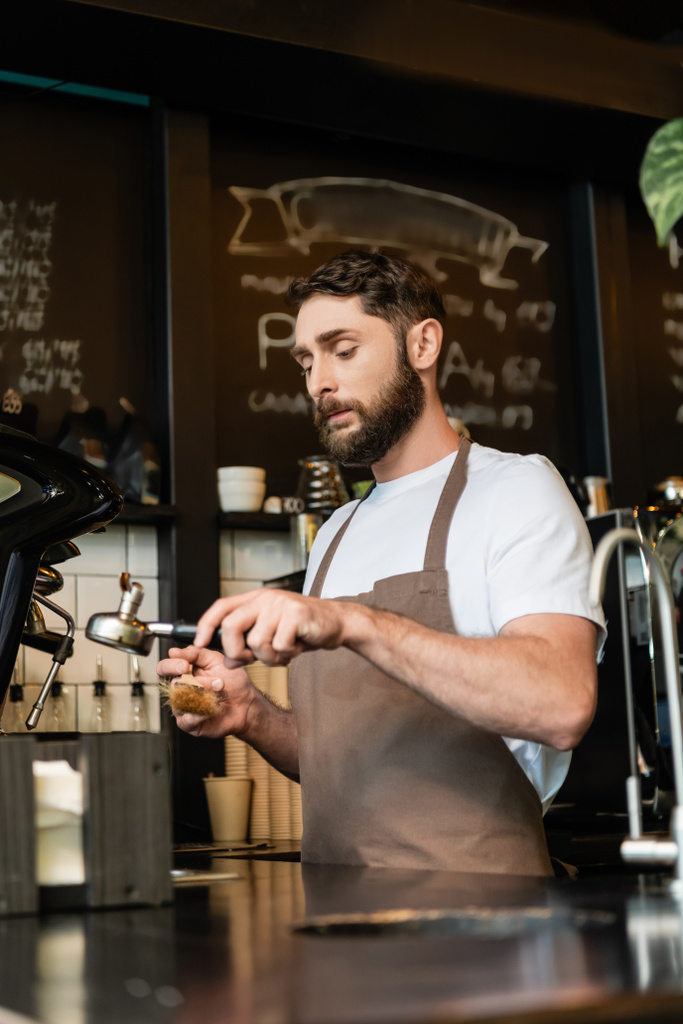 コーヒーショップでブラシが付いているエプロンのクリーニングのコーヒーマシンのホールダーのひげ付きのバリスタ - 写真・画像