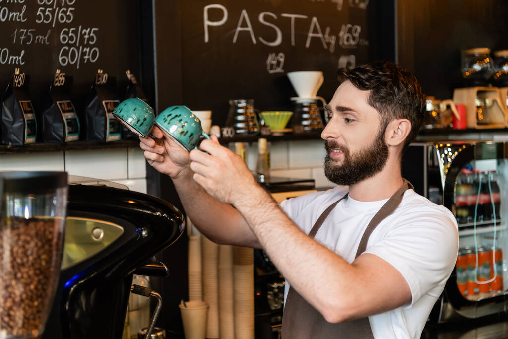 uśmiechnięty brodaty barista w fartuchu trzymający kubki w pobliżu ekspresu do kawy podczas pracy w kawiarni - Zdjęcie, obraz