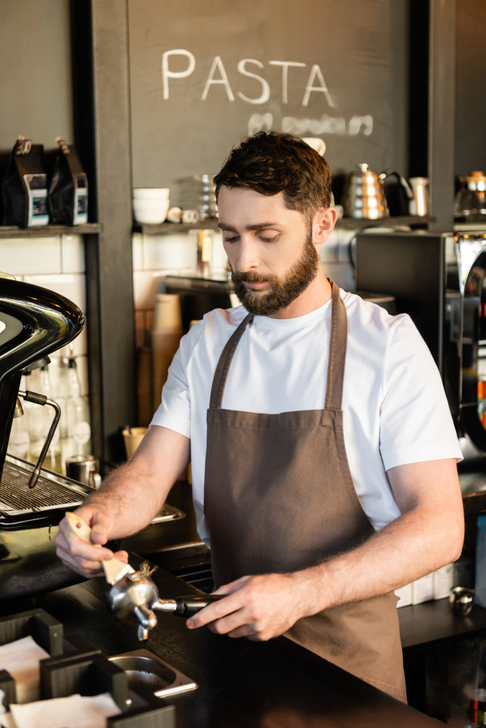 vousatý barista v zástěře čistící držák s kartáčem u kávovaru při práci v kavárně - Fotografie, Obrázek