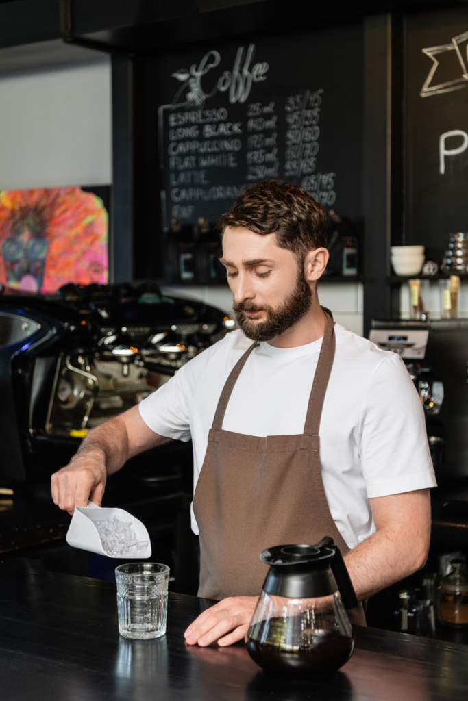 barista in apron pouring ice cubes in glass near coffee pot on bar in coffee shop - Photo, Image