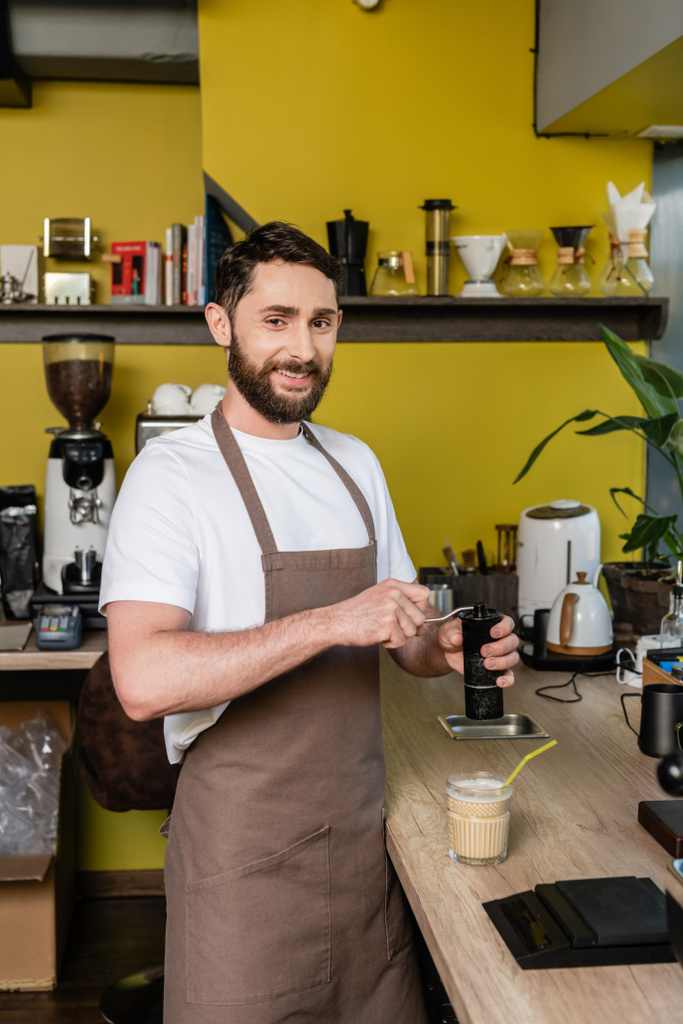 cheerful barista in apron grinding coffee and looking at camera near cold coffee drink in cafe - Photo, Image