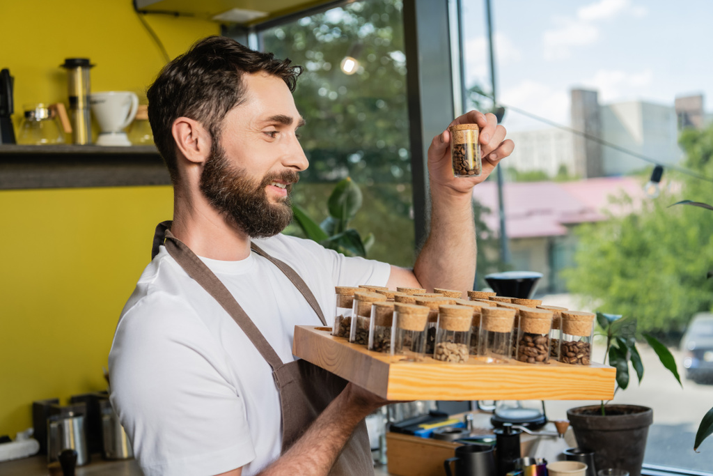 positive bearded barista holding jars with coffee beans while working in coffee shop - Photo, Image