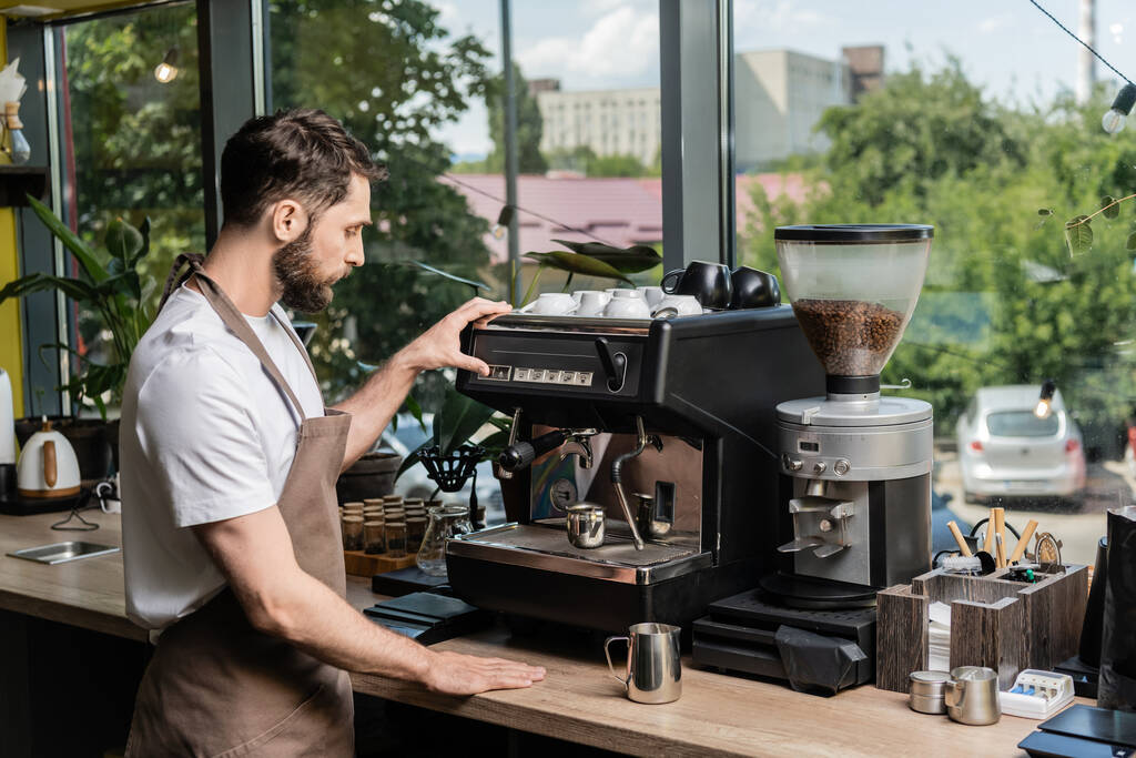 vista laterale del barista barbuto in grembiule fare il caffè sulla macchina da caffè vicino brocche in caffè - Foto, immagini