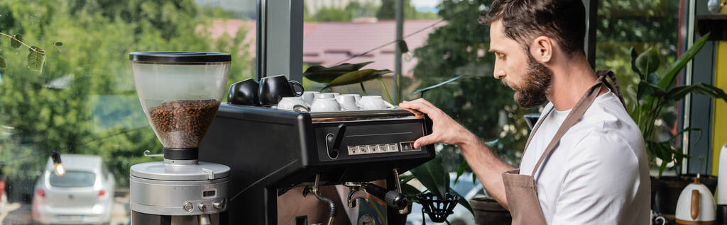 vista lateral de barista barbudo em avental usando máquina de café enquanto trabalhava em café, banner - Foto, Imagem
