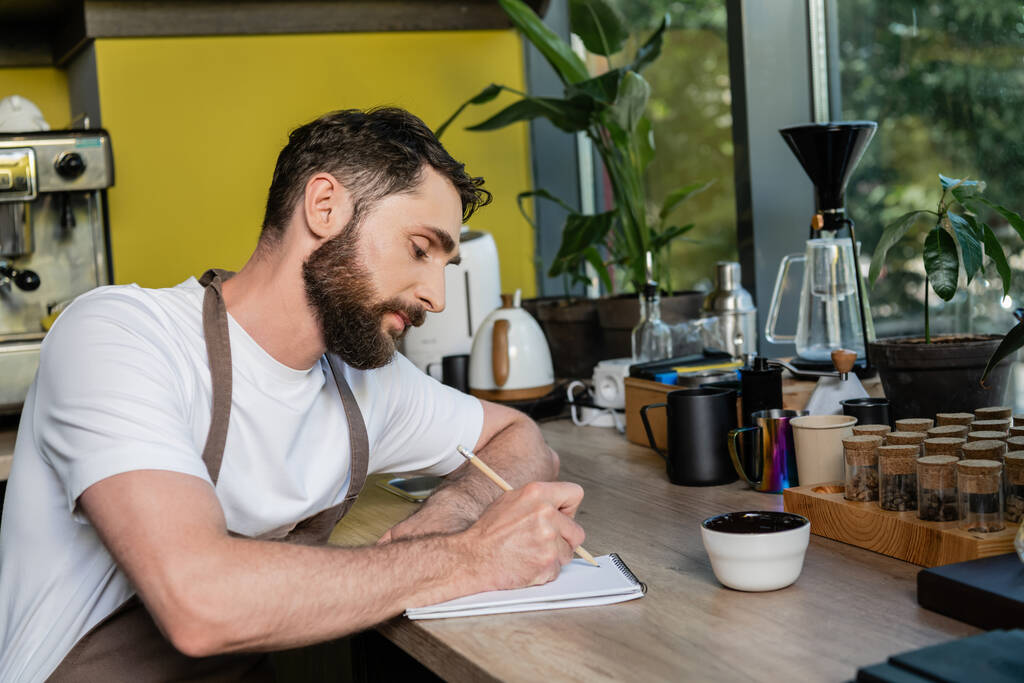 bearded barista in apron writing on notebook near cup and coffee beans in coffee shop - Photo, Image